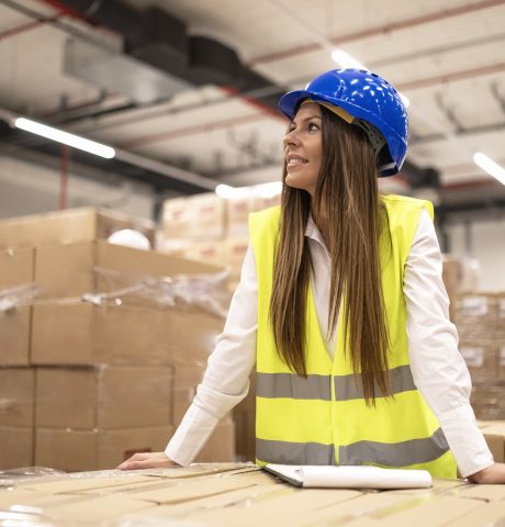 Hardworking professional female worker or manager with hardhat and reflective jacket leaned on cardboard boxes looking aside in large warehouse.